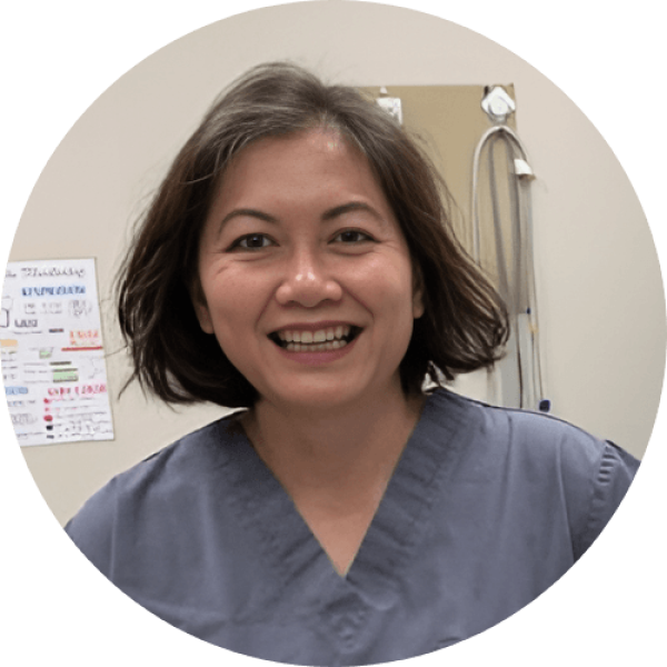 Smiling person in scrubs, in a medical setting with equipment visible in the background.