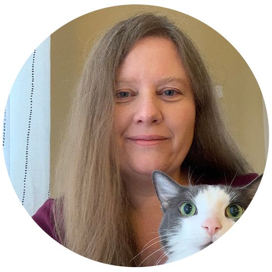 A woman with long hair smiles at the camera, while a gray and white cat with wide eyes sits in front of her.