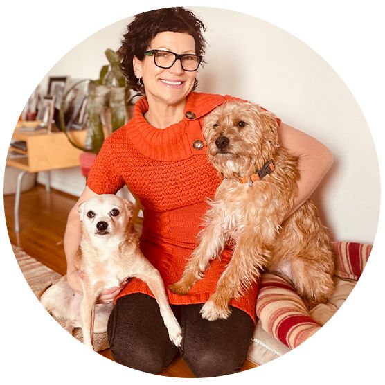 Smiling person in an orange sweater holds two small dogs, one on each side, sitting indoors.