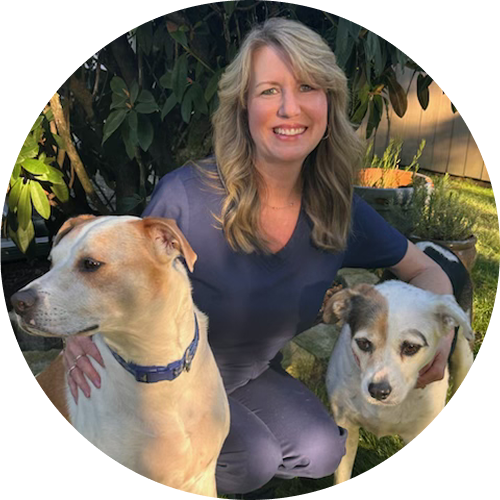 A woman kneels outdoors with two dogs, smiling, as she holds them close.