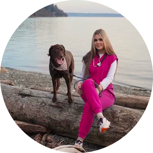 Woman in pink scrubs sitting on driftwood by a beach with a brown dog standing beside her, overlooking the water.