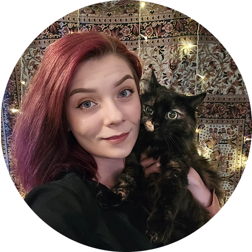 Person with red hair holding a fluffy black cat in front of a patterned background with fairy lights.